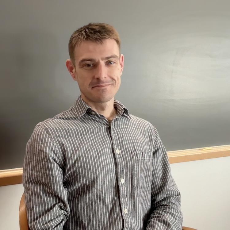 Postdoc, Zach Maschman, sitting in front of a blackboard in a classroom